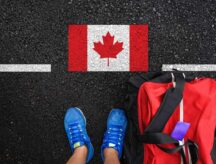 A person with a packed bag stands on a road with the Canadian flag in front of them.