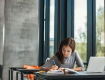 A female student sits at a desk and writes in a big book