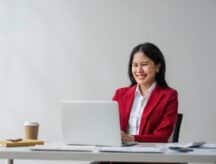 A woman in a red blazer smiles while sitting in front of a laptop.