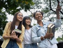 Three international students walk together while carrying books and smiling.