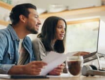 A man and a woman stare at a laptop screen while holding paperwork